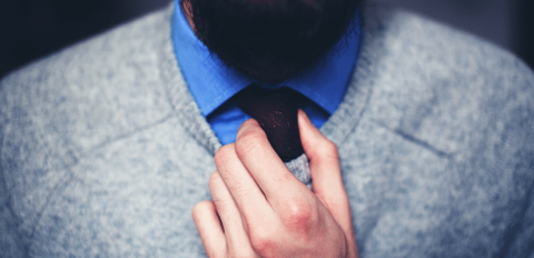 Ein bärtiger Mann zieht unter dem V-Ausschnitt seines Pullovers die Krawatte zurecht